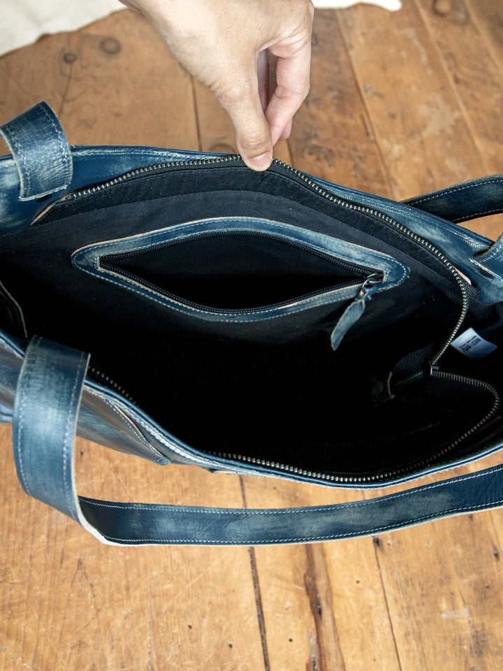 A hand holds open the Tallmadge Leather Handbag by Savannah Sevens western life{&}style, showcasing its blue leather exterior with a zippered inner pocket and a spacious black fabric-lined interior, all set against a wooden floor. This elegant handbag seamlessly blends style and functionality.