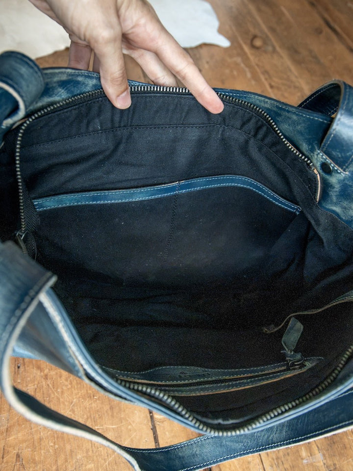 A hand holds open the Savannah Sevens western life{&}style Tallmadge Leather Handbag in blue, revealing its spacious black fabric interior with several pockets and a zipper compartment, all set on a wooden floor.