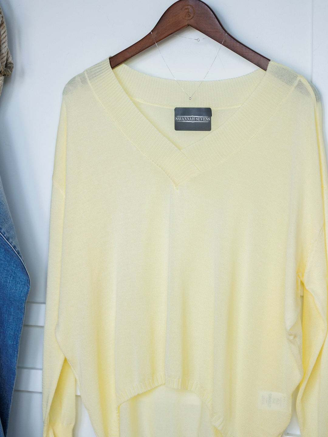 The Tallula Butter Yellow Modal Top by Savannah Sevens western life{&}style, a relaxed fit long sleeve with a black label on the collar, hangs on a wooden hanger against a white background. Partially visible denim appears to the left.