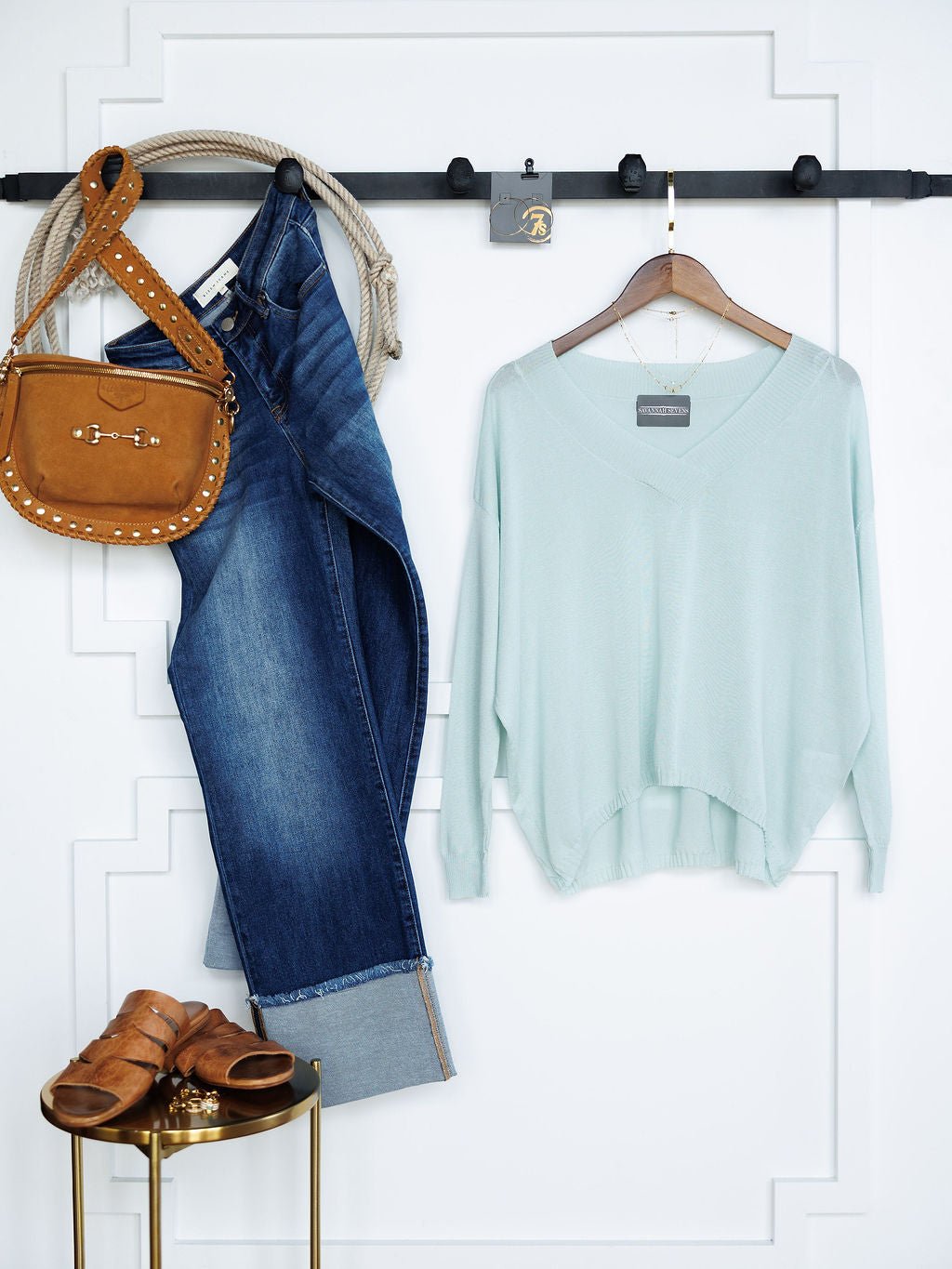 The Tallula Light Seafoam Modal Top by Savannah Sevens western life{&}style hangs on a wooden hanger next to cuffed blue jeans, a studded tan crossbody bag, and tan sandals, all displayed against a white wall with decorative trim.