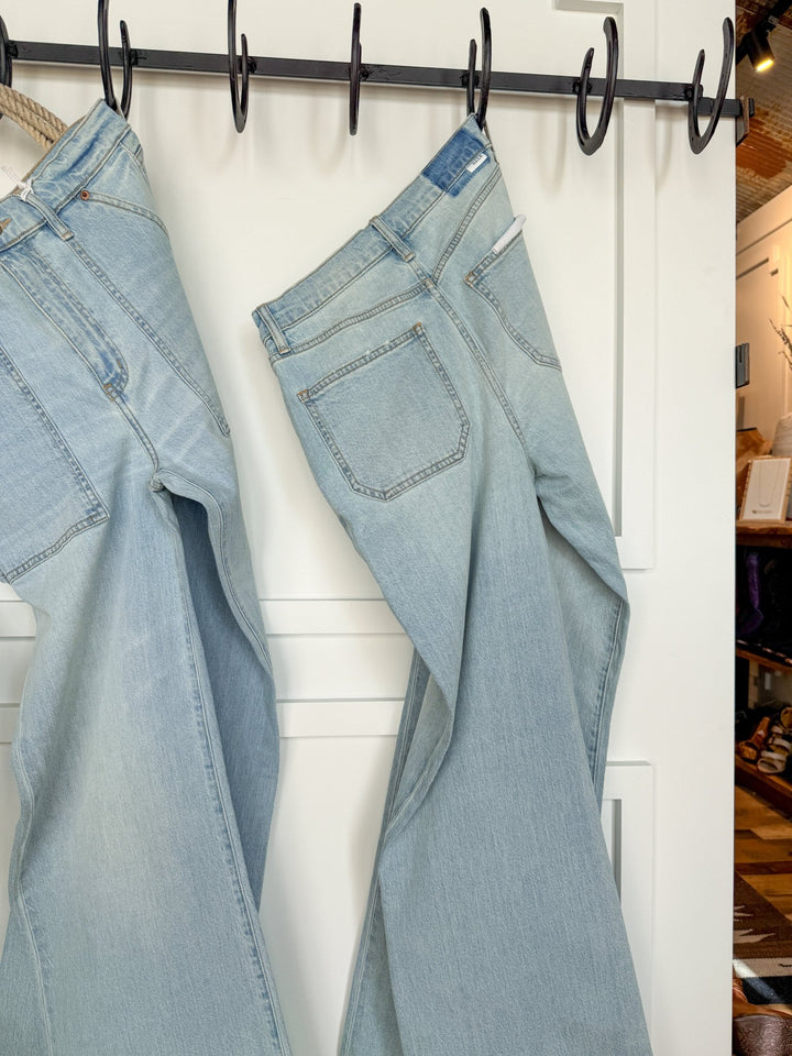 Two pairs of Savannah Sevens western life{&}style The Tallyn High Rise 90's Wide Leg Jeans in light blue hang by their belt loops from black hooks on a white wall, with part of a store interior visible to the right.