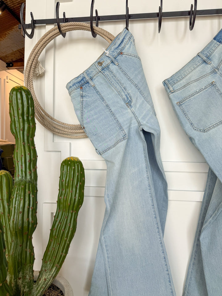 The Tallyn High Rise 90's Wide Leg Jean by Savannah Sevens western life{&}style in light blue hangs on a hook beside a coiled rope and a potted green cactus, set against a white paneled wall.