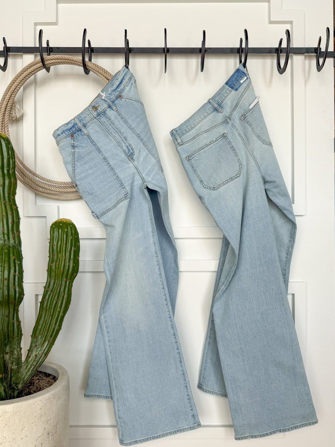 Two pairs of The Tallyn High Rise 90's Wide Leg Jean by Savannah Sevens western life{&}style hang in light blue on black hooks against a white wall, with a potted cactus on the left and rope decor draped over the hooks.