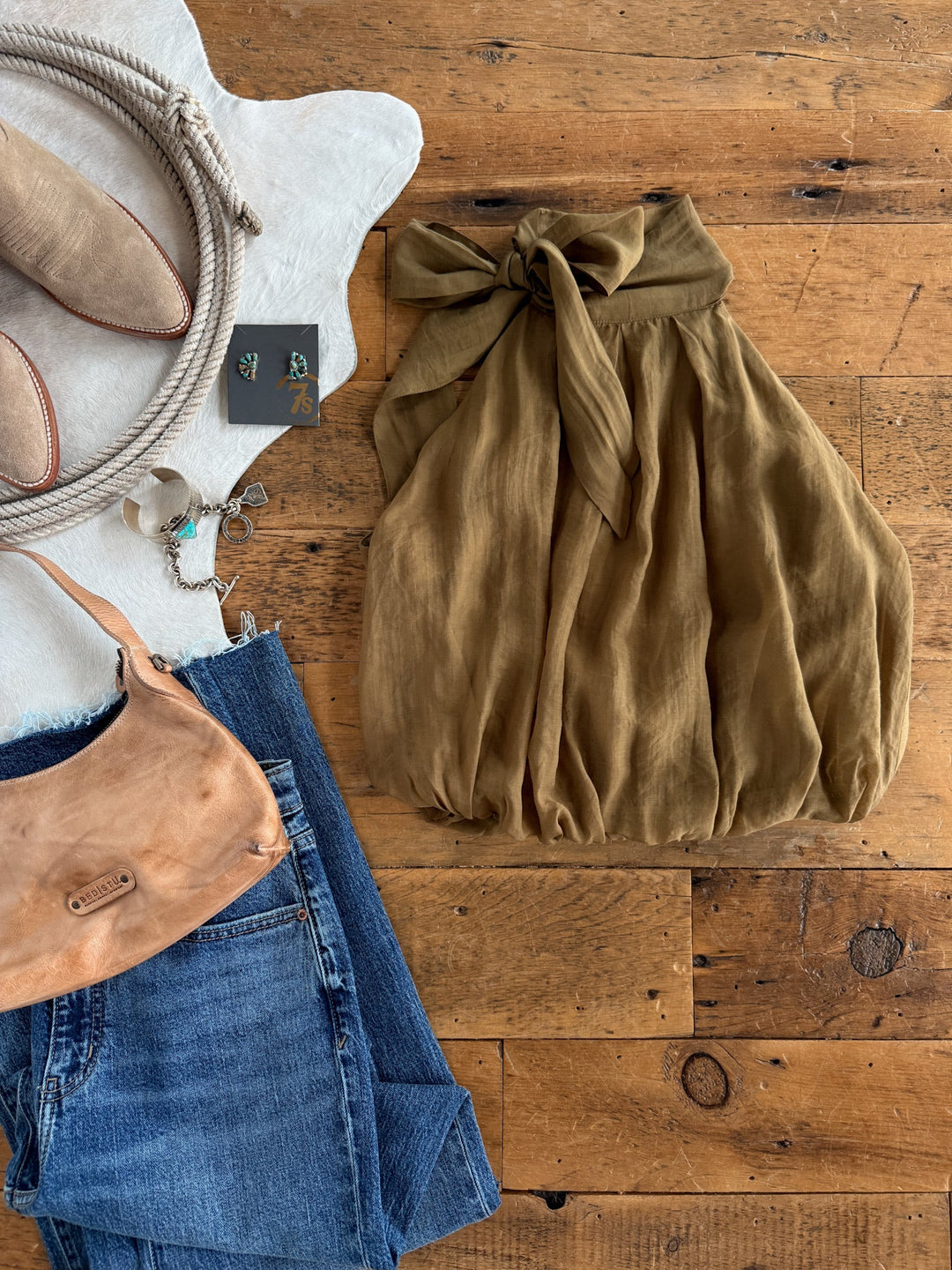 A flat lay featuring the Talmage Olive Tencel Halter Neck Top by Savannah Sevens western life{&}style, paired with blue jeans, a tan purse, tan boots, and jewelry on a wooden floor.