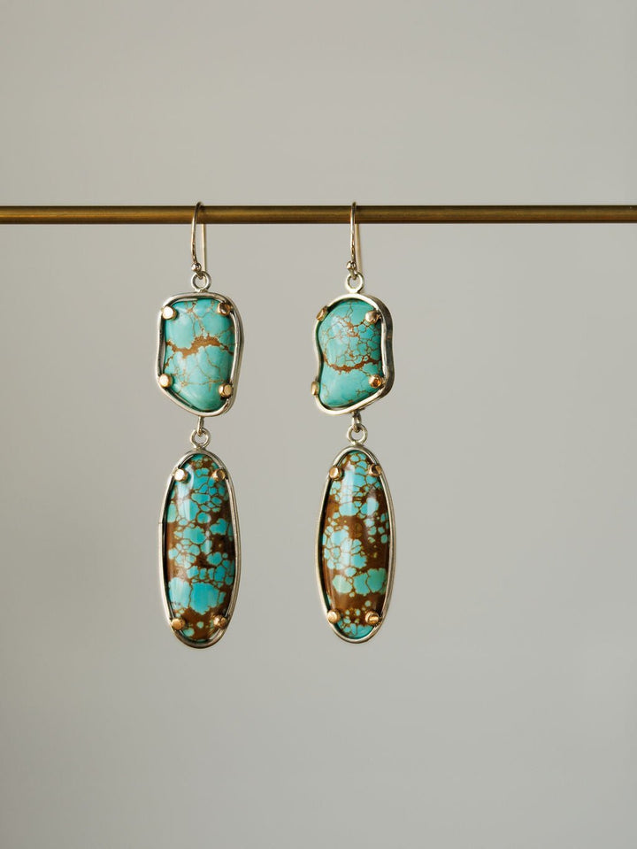 The Taos Earrings by Savannah Sevens western life{&}style feature sterling silver and irregular Number 8 turquoise stones with brown and gold matrix, set in silver frames and displayed against a plain background.
