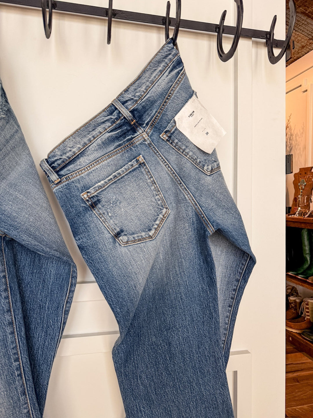 The Taryn High Rise Straight Leg Jean by Savannah Sevens western life{&}style hangs by the waistband on a black hook rack against a white panel wall with a white tag on the back pocket. Another pair of jeans is partially visible on the left.