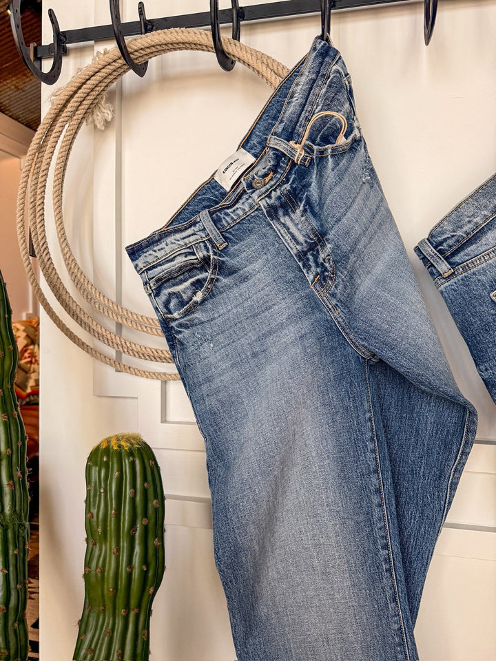 The Taryn High Rise Straight Leg Jean by Savannah Sevens western life{&}style hangs on a black hook in front of a white wall, with a coiled rope and green cactus nearby.