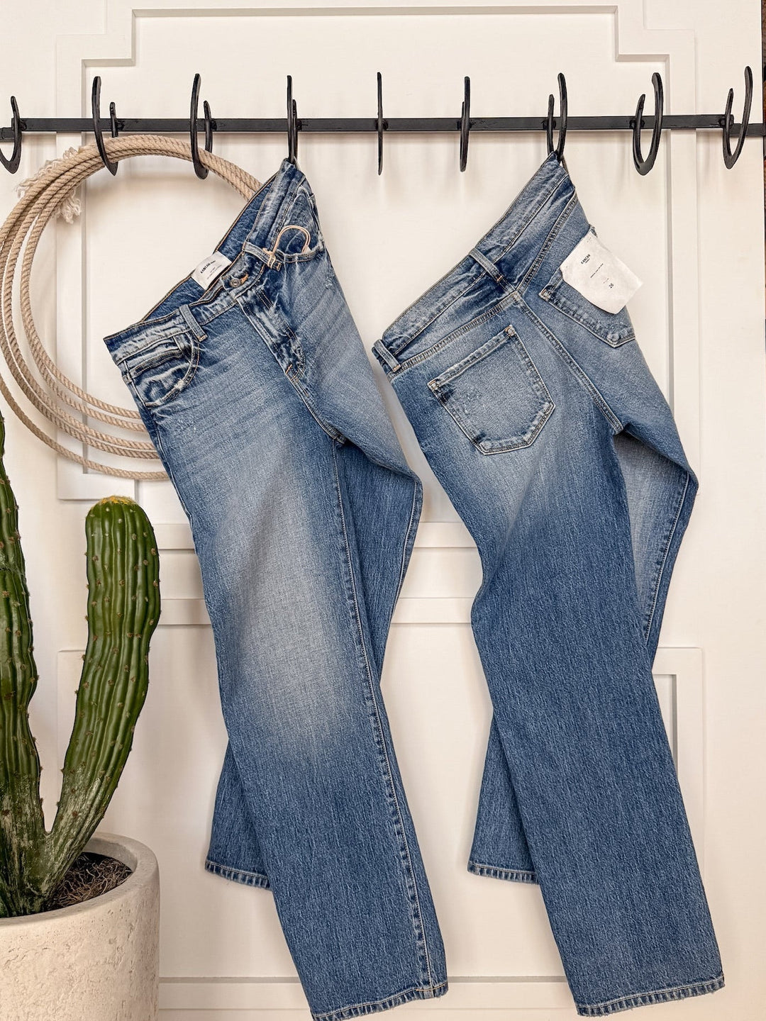 Two pairs of The Taryn High Rise Straight Leg Jeans by Savannah Sevens western life{&}style in medium wash hang side by side on black hooks against a white wall, next to a green potted cactus and a coiled rope.