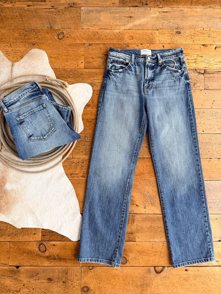 A pair of medium wash denim jeans is laid flat on a wooden floor next to a folded Savannah Sevens western life{&}style The Taryn High Rise Straight Leg Jean, placed on a coiled rope and a light cowhide rug.