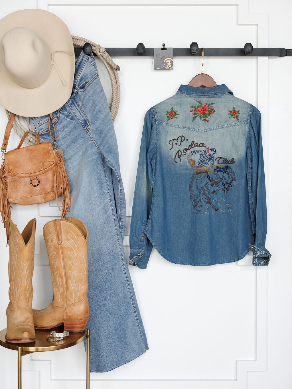 A Western-themed outfit hangs on the wall: a Savannah Sevens western life{&}style Tasha Rodeo Club Embroidered Denim Button Up, a cowboy hat, denim jeans, tan boots, and a brown fringe purse.
