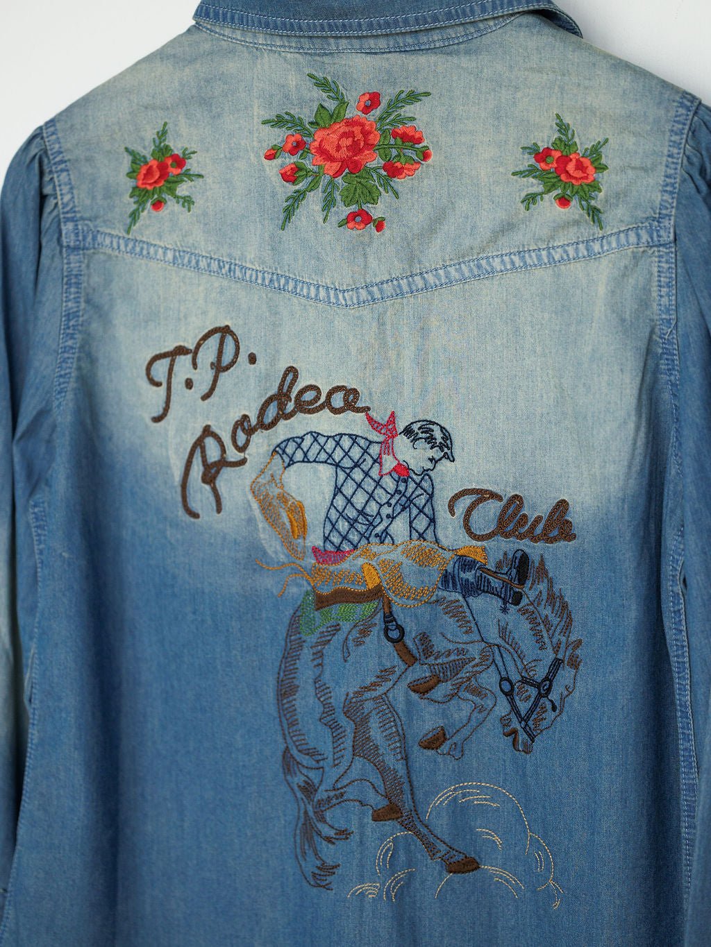 The Tasha Rodeo Club Embroidered Denim Button Up by Savannah Sevens western life{&}style features red flower embroidery on the shoulders and a cowboy with "T.P. Rodeo Club" on the back—a signature Tasha Polizzi touch.