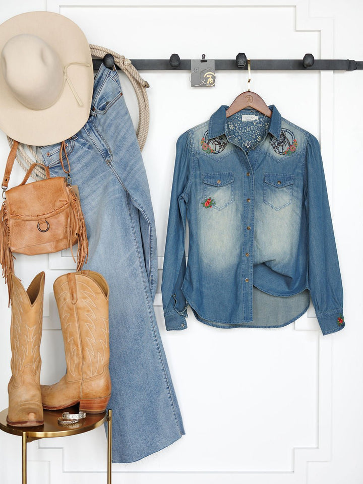 A beige hat, rope, blue jeans, tan purse, tan cowboy boots, and a Tasha Rodeo Club Embroidered Denim Button Up by Savannah Sevens western life{&}style are neatly displayed on a wall rack and stool against a white wall.