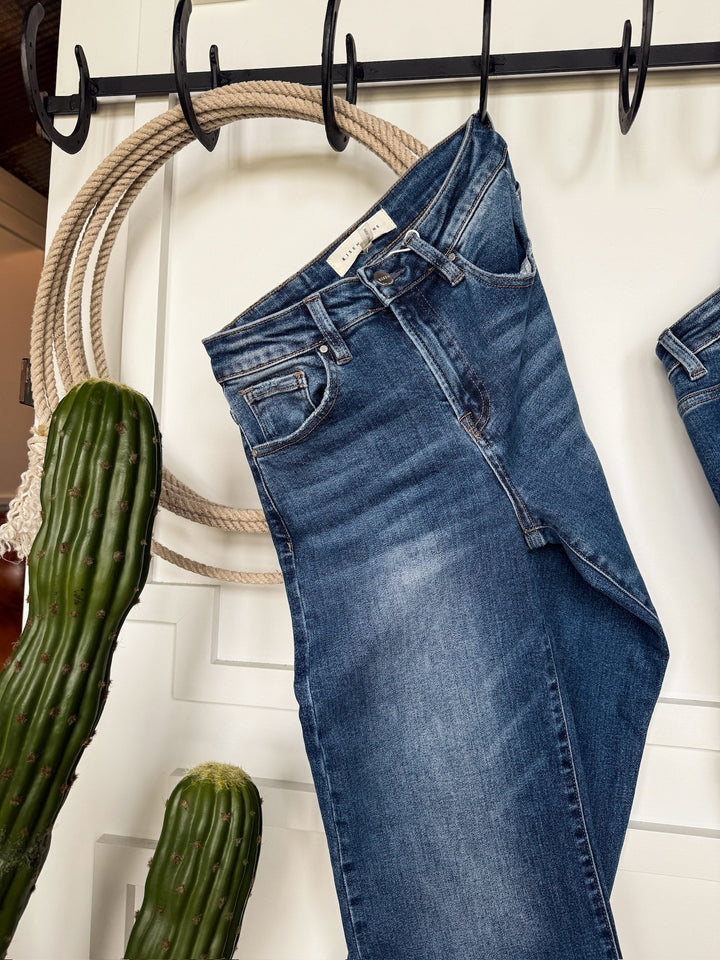 The Tatum High Rise Relaxed Straight Leg Jean by Savannah Sevens western life{&}style in medium wash hangs on a black hook against a white wall, with two tall green cacti nearby.