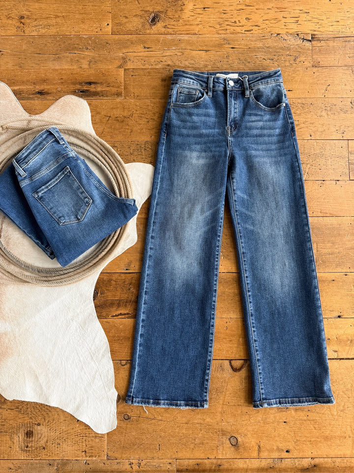 Two pairs of The Tatum High Rise Relaxed Straight Leg Jean by Savannah Sevens western life{&}style are shown: one folded on a light cowhide rug, the other ultra high rise pair laid flat beside it on a wooden floor.