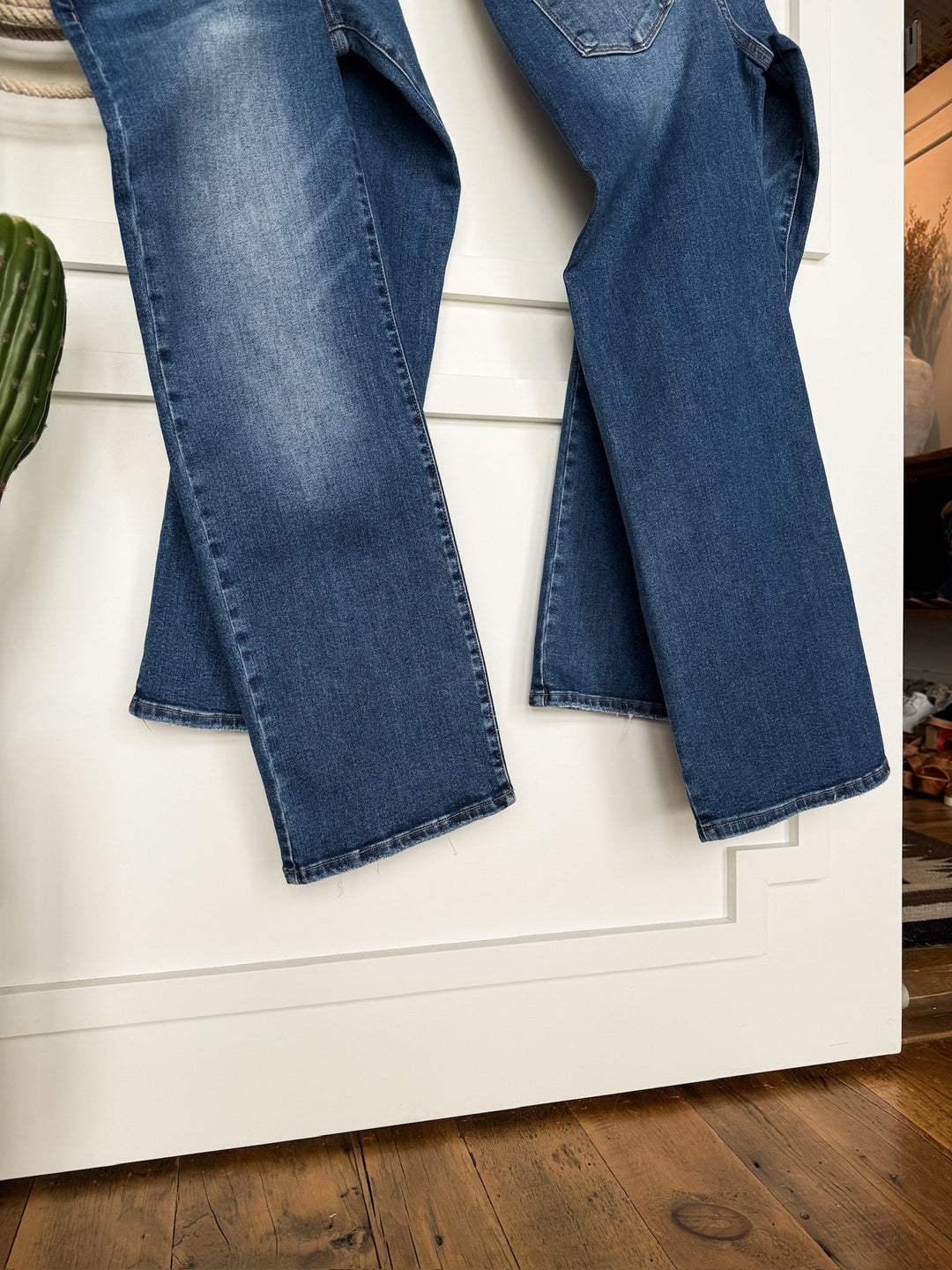 The Tatum High Rise Relaxed Straight Leg Jean by Savannah Sevens western life{&}style in medium wash denim hangs on a white door with decorative trim, with a wooden floor and a green plant visible in the foreground.