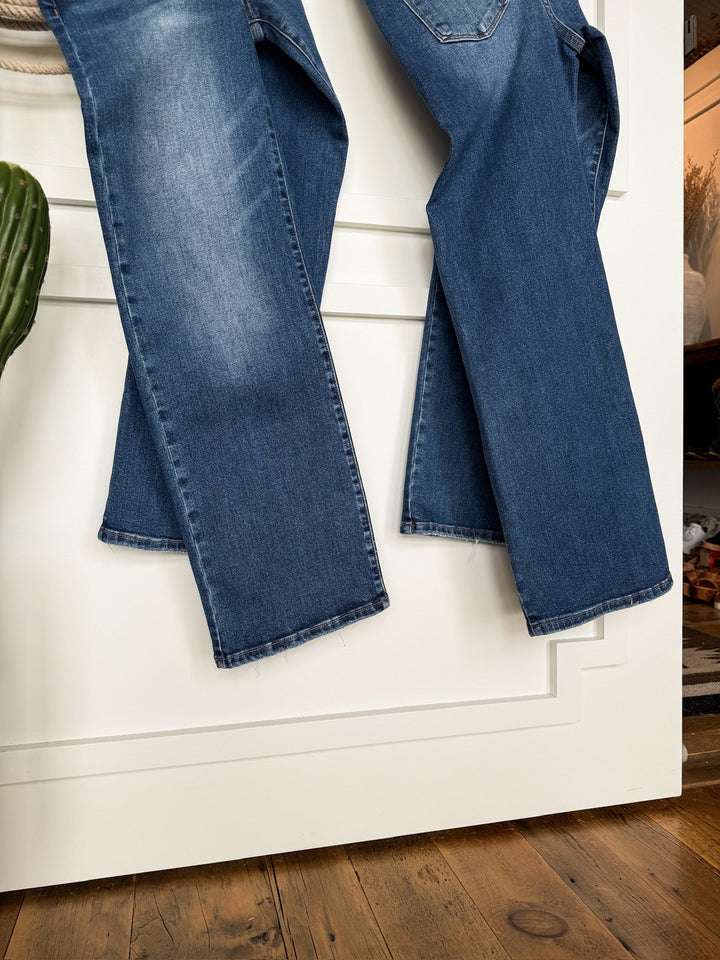 The Tatum High Rise Relaxed Straight Leg Jean by Savannah Sevens western life{&}style in medium wash denim hangs on a white door with decorative trim, with a wooden floor and a green plant visible in the foreground.