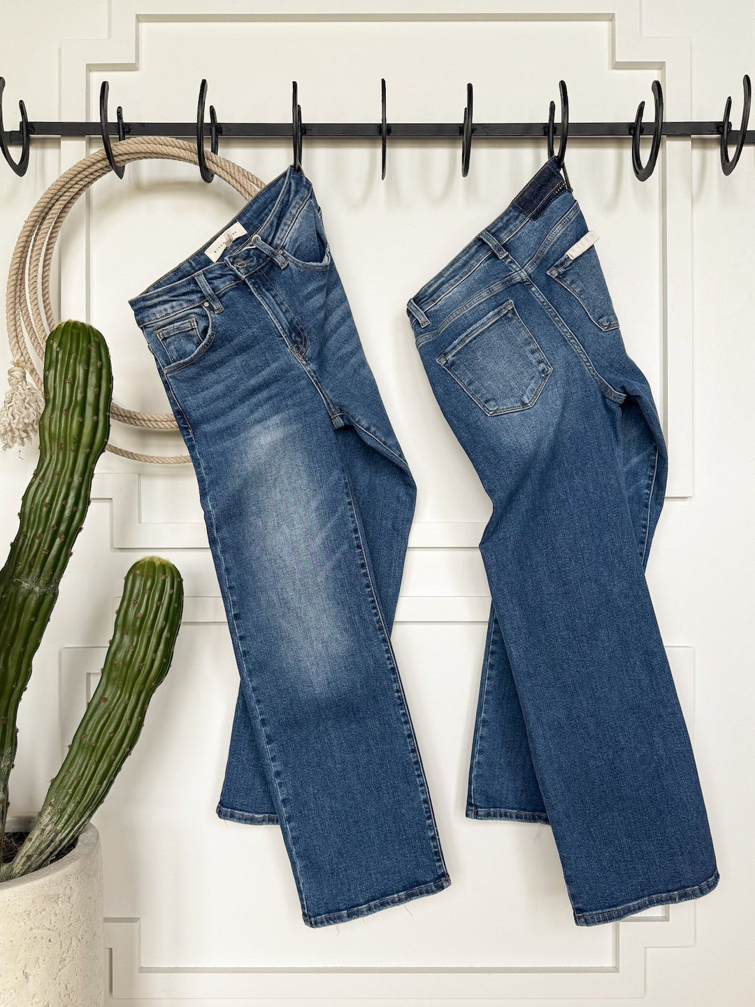 Two pairs of Savannah Sevens western life{&}style's The Tatum High Rise Relaxed Straight Leg Jeans in medium wash hang side by side on black hooks against a white paneled wall, with a potted cactus and coiled rope to the left.