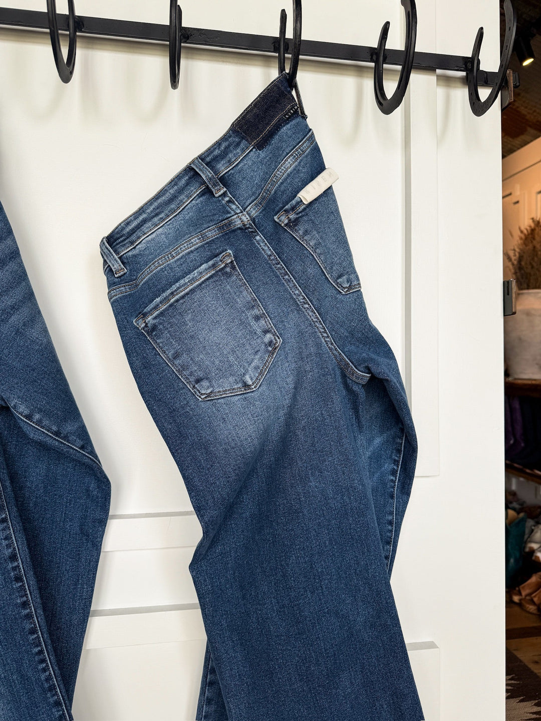 The Tatum High Rise Relaxed Straight Leg Jean by Savannah Sevens western life{&}style, in a medium wash, hangs on a black hooked rack against a white door with a white tag visible on the waistband.