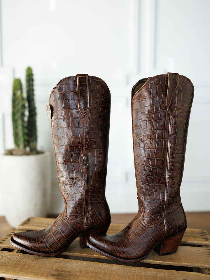 The Tavares by Savannah Sevens western life{&}style: brown, knee-high snip toe boots with a crocodile pattern in exotic leather, side zippers, shown on wood with a potted cactus in the background. Boots by Ariat. 