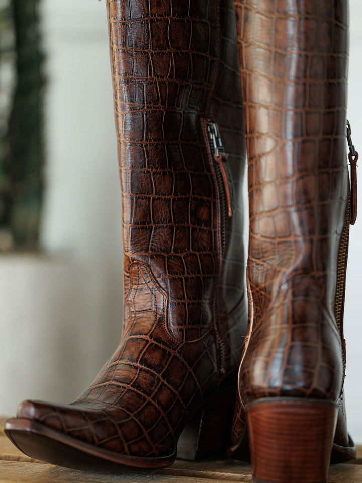 Close-up of The Tavares by Savannah Sevens western life{&}style: brown, knee-high, crocodile-embossed leather snip toe boots with wooden block heels and side zippers, displayed indoors with a blurred plant in the background. Boots by Ariat. 