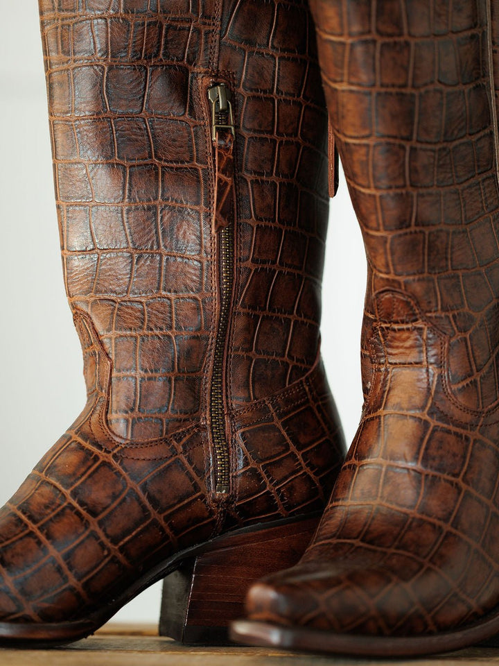 A close-up of The Tavares cowboy boots by Savannah Sevens western life{&}style, featuring brown exotic leather with a crocodile pattern, side zipper, wooden heels, and a snip toe design. Boots by Ariat.