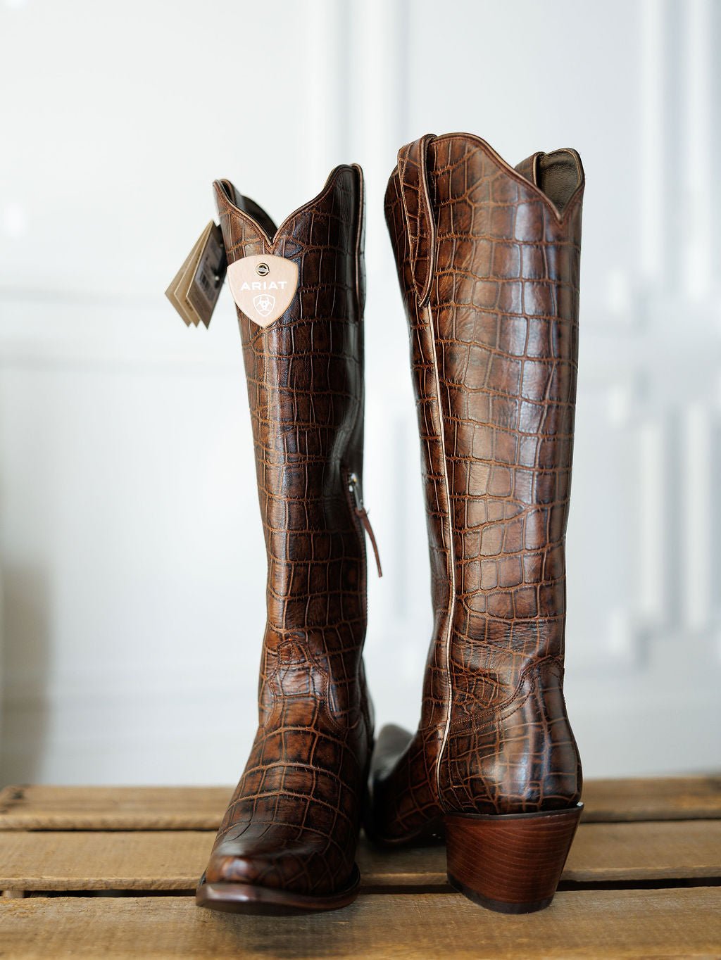The Tavares by Savannah Sevens western life{&}style: tall brown exotic leather cowboy boots with a crocodile pattern and snip toes, standing upright on wood against a blurred white background. Boots by Ariat.