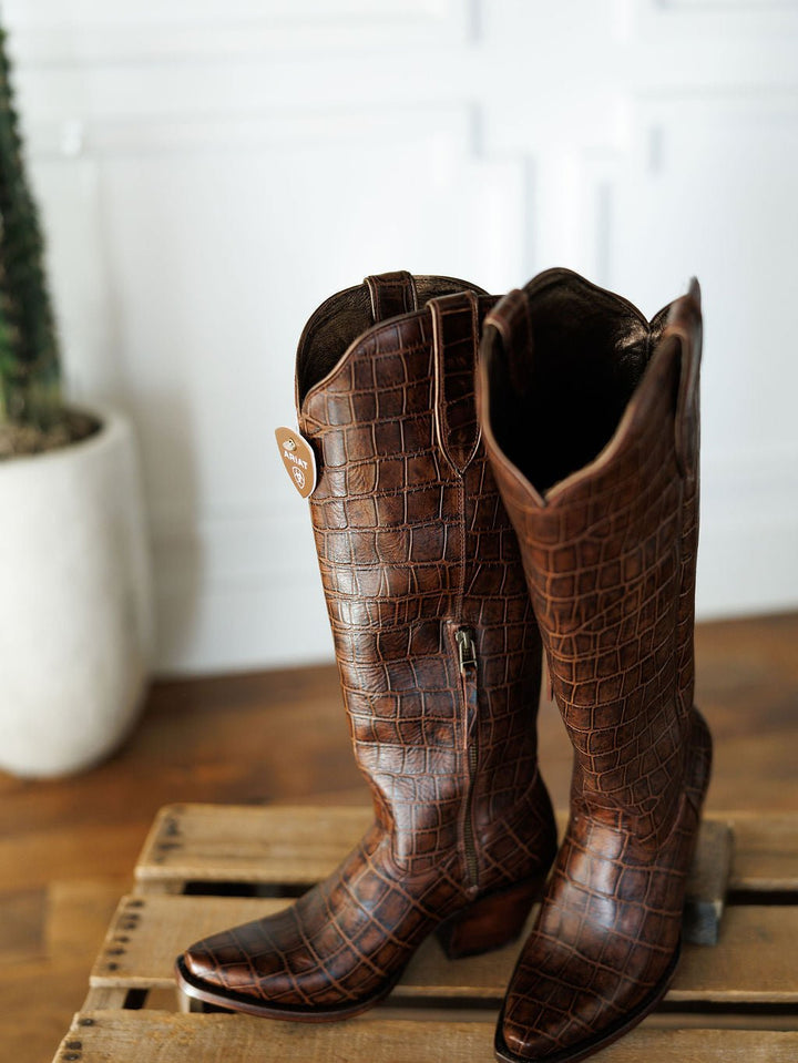 A pair of tall, brown "The Tavares" snip toe boots by Savannah Sevens western life{&}style with a crocodile pattern sit on a wooden crate. A potted cactus is in the background against a white paneled wall. Boots by Ariat.