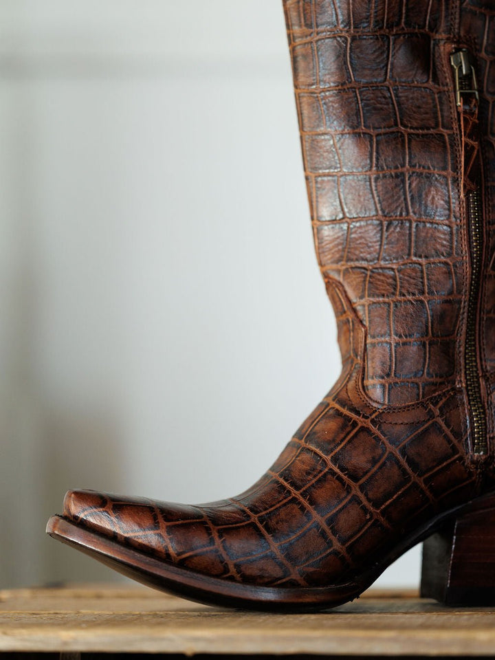 A close-up of The Tavares by Savannah Sevens western life{&}style, a snip toe boot with exotic leather in a crocodile skin pattern and a zipper, resting on a wooden surface. Boots by Ariat.
