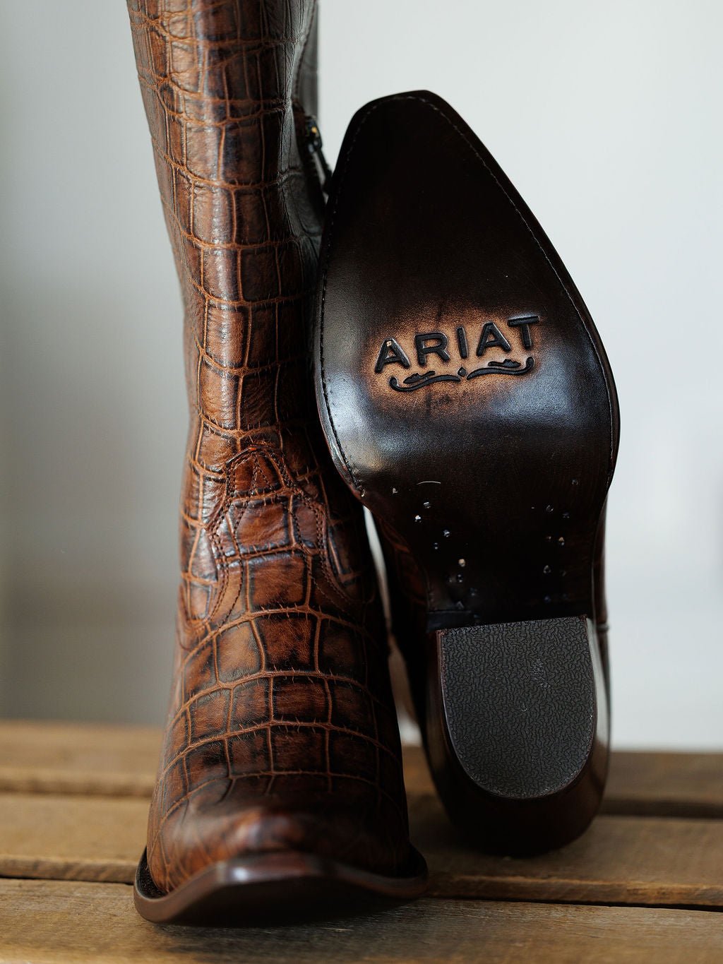 A close-up of The Tavares brown snip toe boot by Savannah Sevens western life{&}style, featuring exotic crocodile-patterned leather, sits on a wooden surface next to another boot showing its sole. Boots by Ariat. 