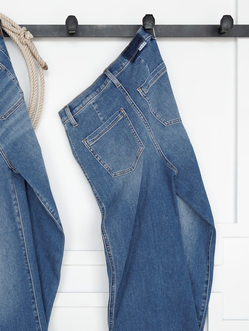 The Taylee High Rise Wide Leg Jean by Savannah Sevens western life{&}style in medium wash denim hangs from a black wall hook by the belt loop, beside another denim piece, against a white wall with decorative trim.