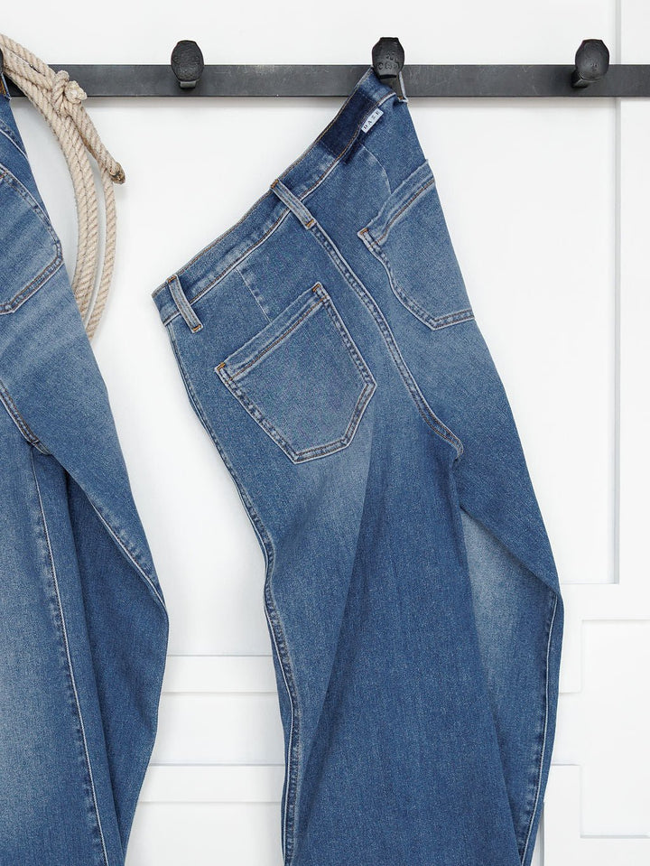 The Taylee High Rise Wide Leg Jean by Savannah Sevens western life{&}style in medium wash denim hangs from a black wall hook by the belt loop, beside another denim piece, against a white wall with decorative trim.