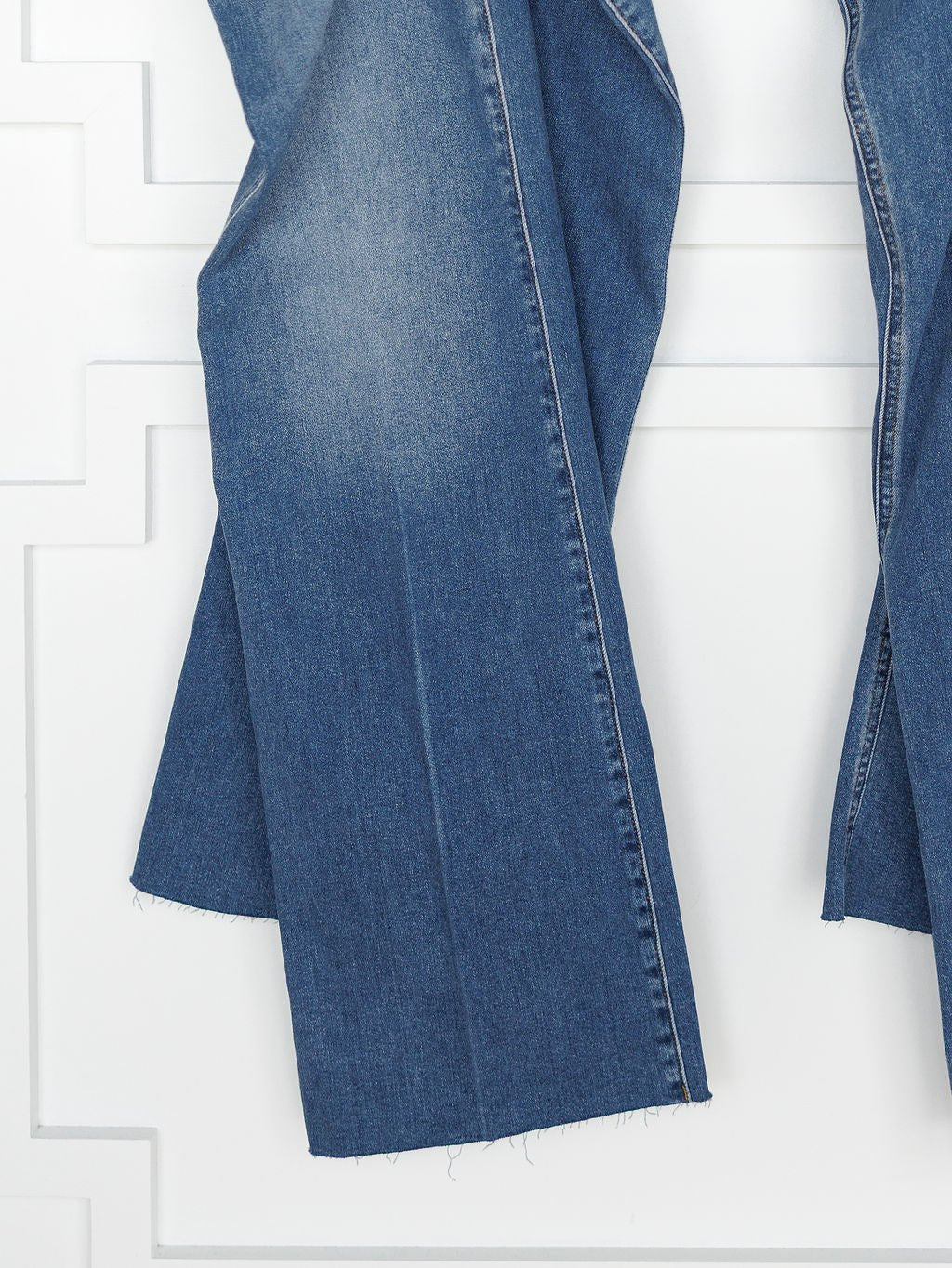 A close-up of the lower legs of The Taylee High Rise Wide Leg Jean by Savannah Sevens western life{&}style, in medium wash denim with raw unfinished hems, hanging against a white textured wall.