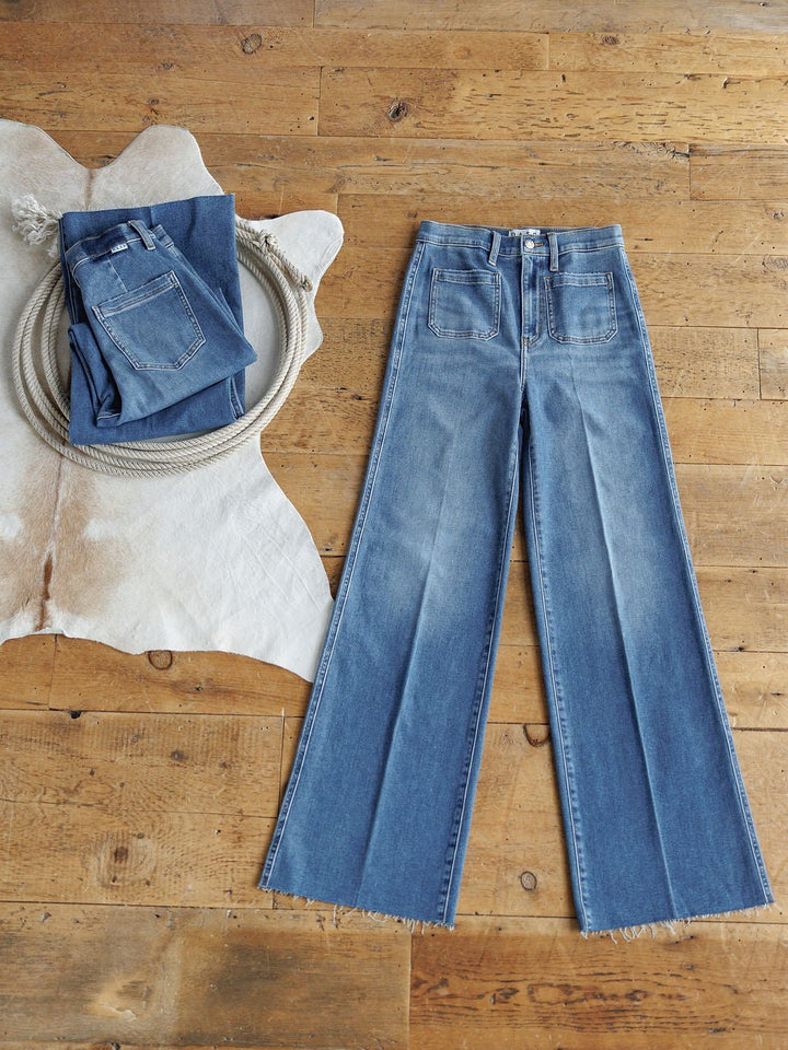 Two pairs of blue Savannah Sevens western life{&}style jeans are on a wooden floor. One, The Taylee High Rise Wide Leg Jean with frayed hems and large front pockets, is laid flat; the other is folded on a cowhide rug next to a coiled rope.