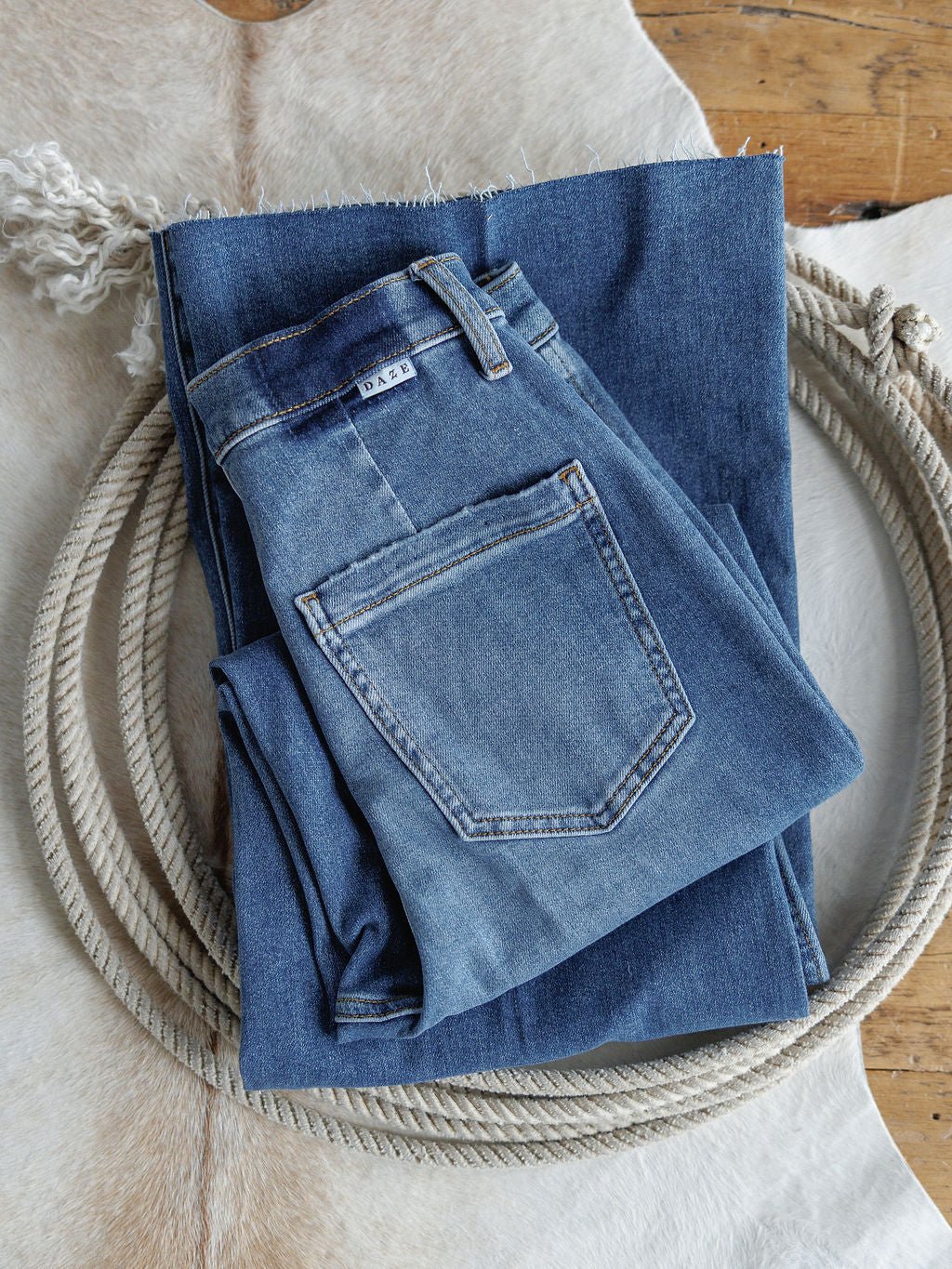 The Taylee High Rise Wide Leg Jean by Savannah Sevens western life{&}style is shown folded on denim fabric, both displayed atop a coiled rope and light cowhide rug on a wooden floor.