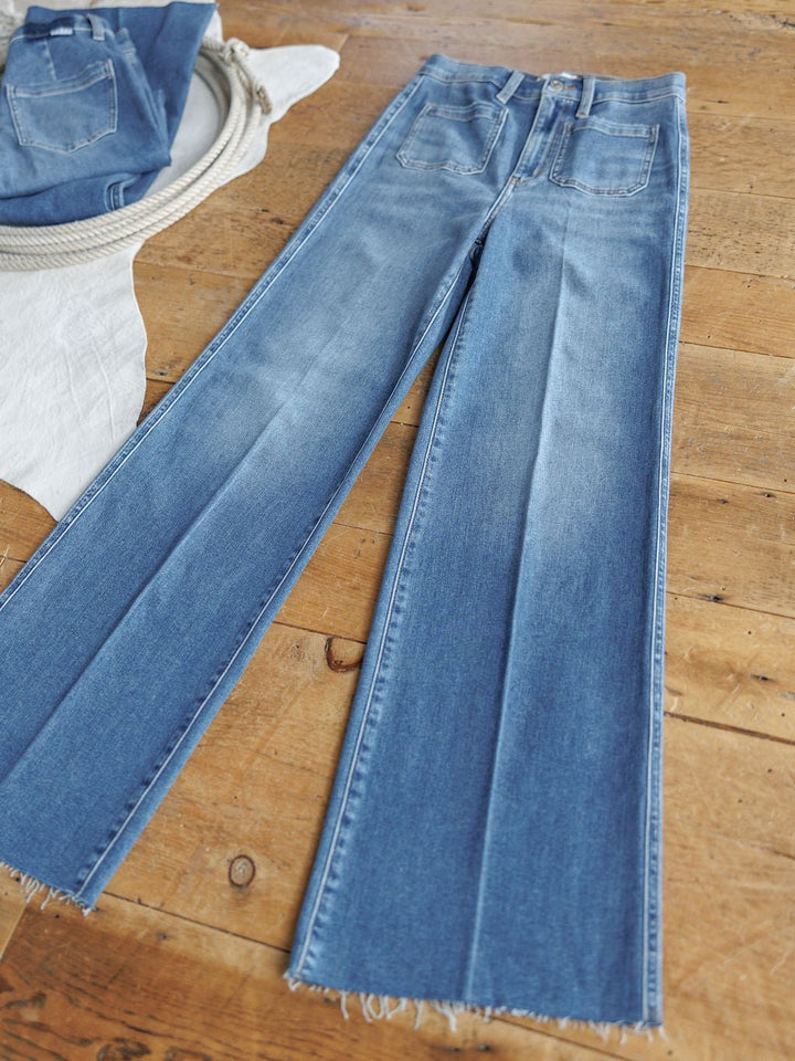 The Taylee High Rise Wide Leg Jean from Savannah Sevens western life{&}style in medium wash denim is displayed flat on a wooden floor with folded jeans, a coiled rope, and a beige cloth nearby.