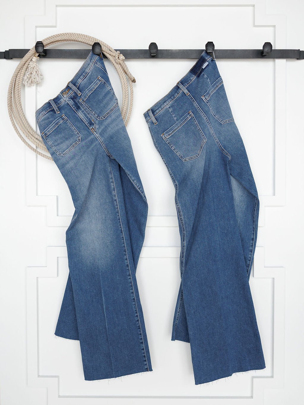Two pairs of The Taylee High Rise Wide Leg Jeans by Savannah Sevens western life{&}style in medium wash hang side by side on a black wall rack. A coiled rope is on the left, against a white wall with decorative trim.