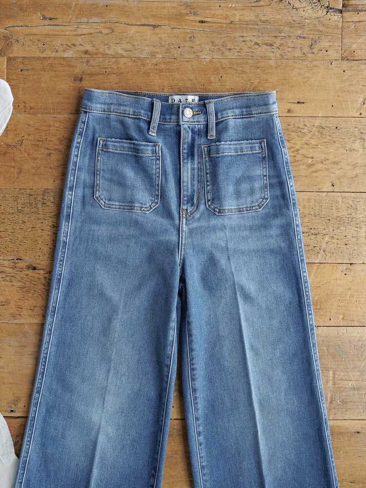 The Taylee High Rise Wide Leg Jean by Savannah Sevens western life{&}style in medium wash denim, featuring a high waist and front patch pockets, is laid flat on a wooden floor.