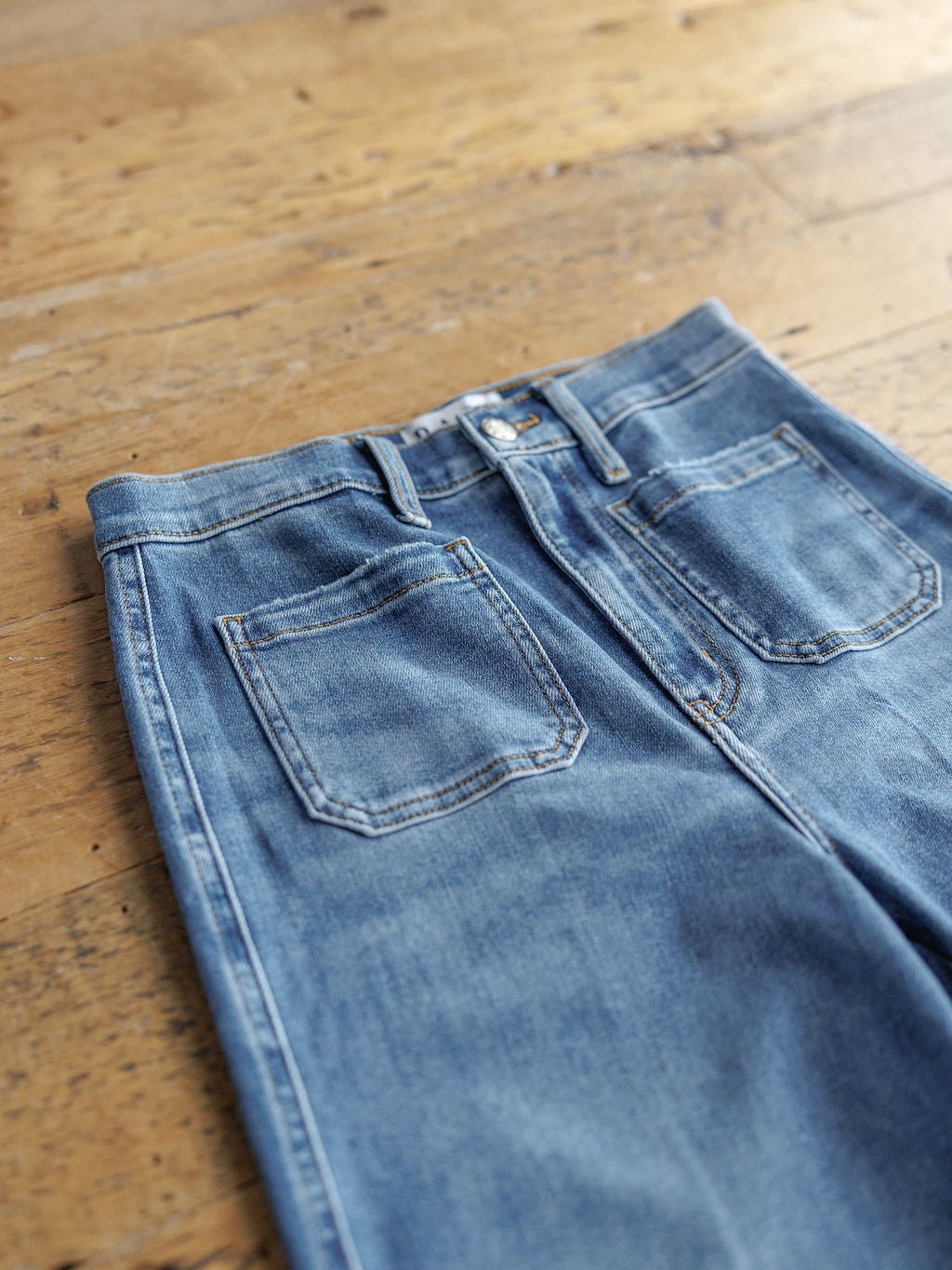 A pair of The Taylee High Rise Wide Leg Jean by Savannah Sevens western life{&}style lies flat on a wooden floor, showcasing medium wash denim with back pockets, visible stitching, and belt loops.