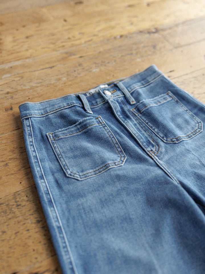 A pair of The Taylee High Rise Wide Leg Jean by Savannah Sevens western life{&}style lies flat on a wooden floor, showcasing medium wash denim with back pockets, visible stitching, and belt loops.