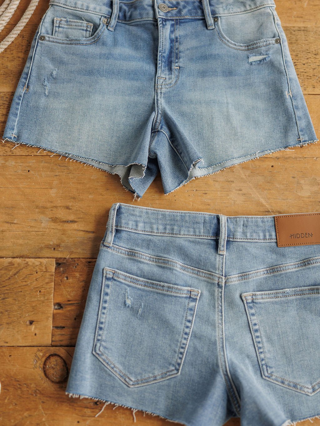 Two pairs of The Taylin Shorts by Savannah Sevens western life{&}style, in a medium light wash with raw cut hems, are displayed flat on wood; one shows the front and the other the back with pockets and a brown leather patch.