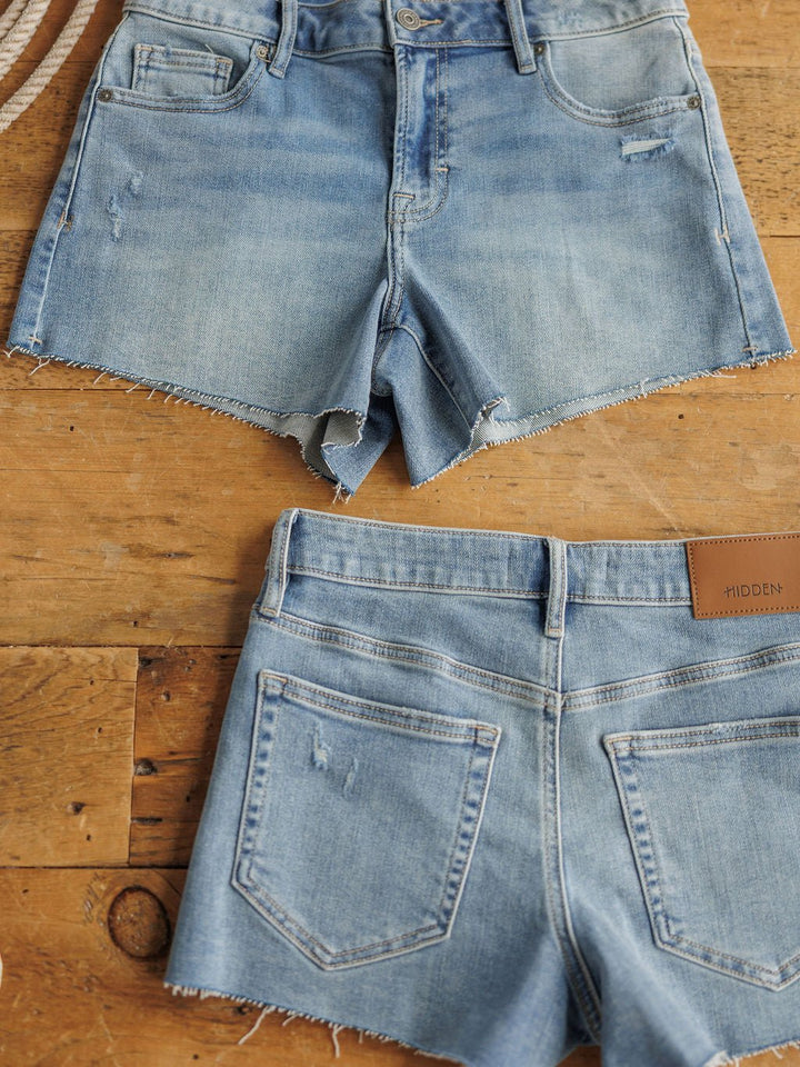 Two pairs of The Taylin Shorts by Savannah Sevens western life{&}style, in a medium light wash with raw cut hems, are displayed flat on wood; one shows the front and the other the back with pockets and a brown leather patch.