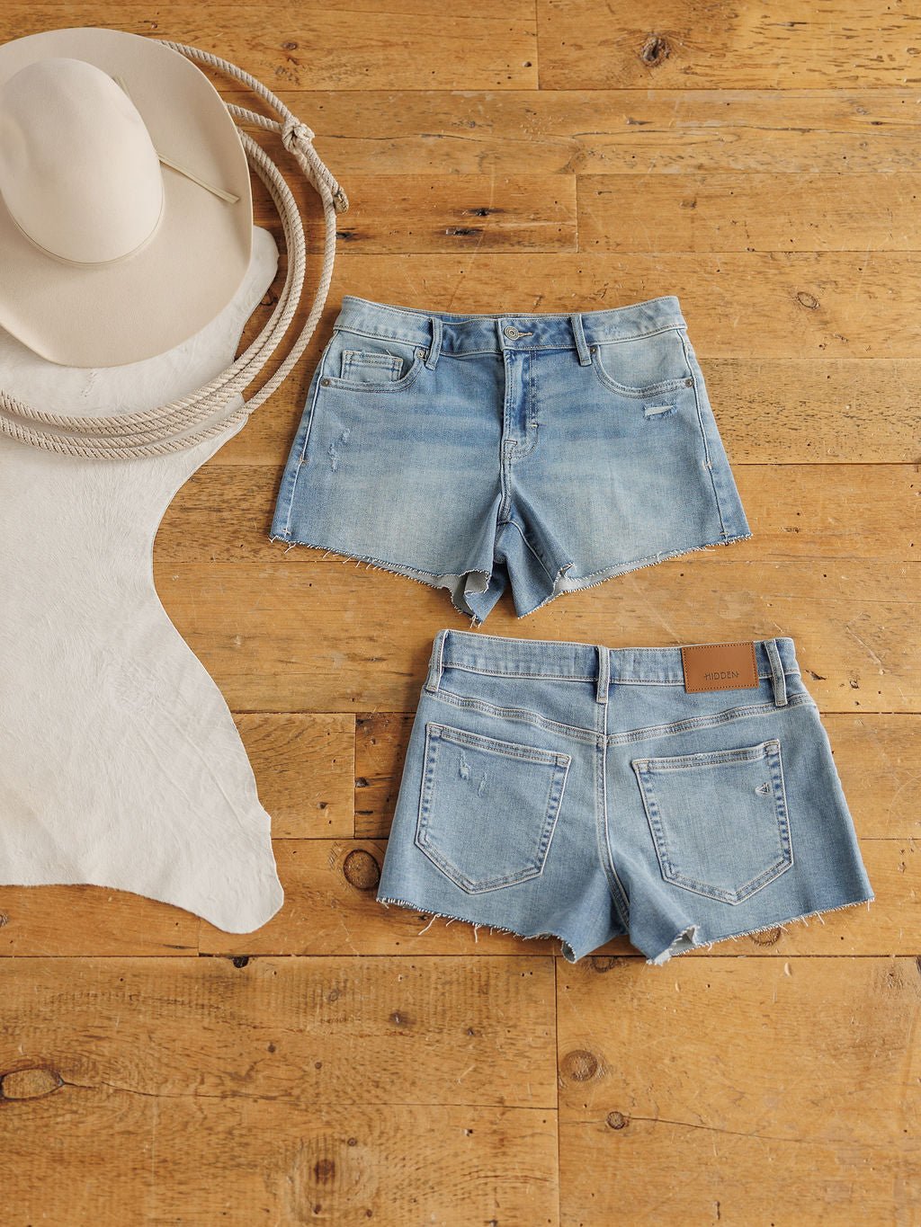 The Taylin Shorts by Savannah Sevens western life{&}style are light blue premier rise denim with a raw cut hem, shown front and back on wood flooring beside a beige cowboy hat, coiled rope, and pale hide.