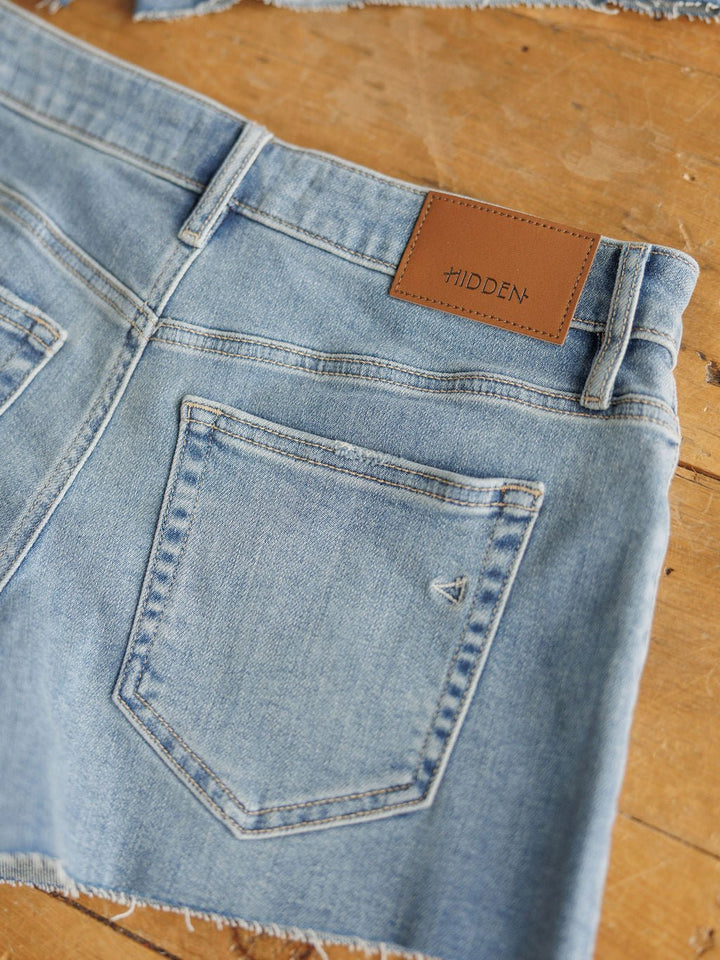 The Taylin Shorts by Savannah Sevens western life{&}style: light blue rise denim shorts with raw cut hems, back pockets, and a brown HIDDEN leather patch on the waistband, shown on a wooden surface.