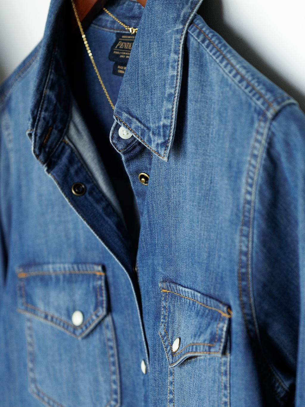 The Tekamah denim shirt by Savannah Sevens western life{&}style, featuring front snap pockets, hangs on a wooden hanger, partially unbuttoned.