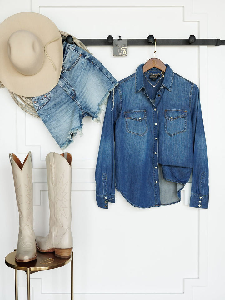 The Tekamah Savannah Sevens western life{&}style hangs on a wall rack besides frayed denim shorts a, with cream cowboy boots on a gold side table in front of a white paneled wall.