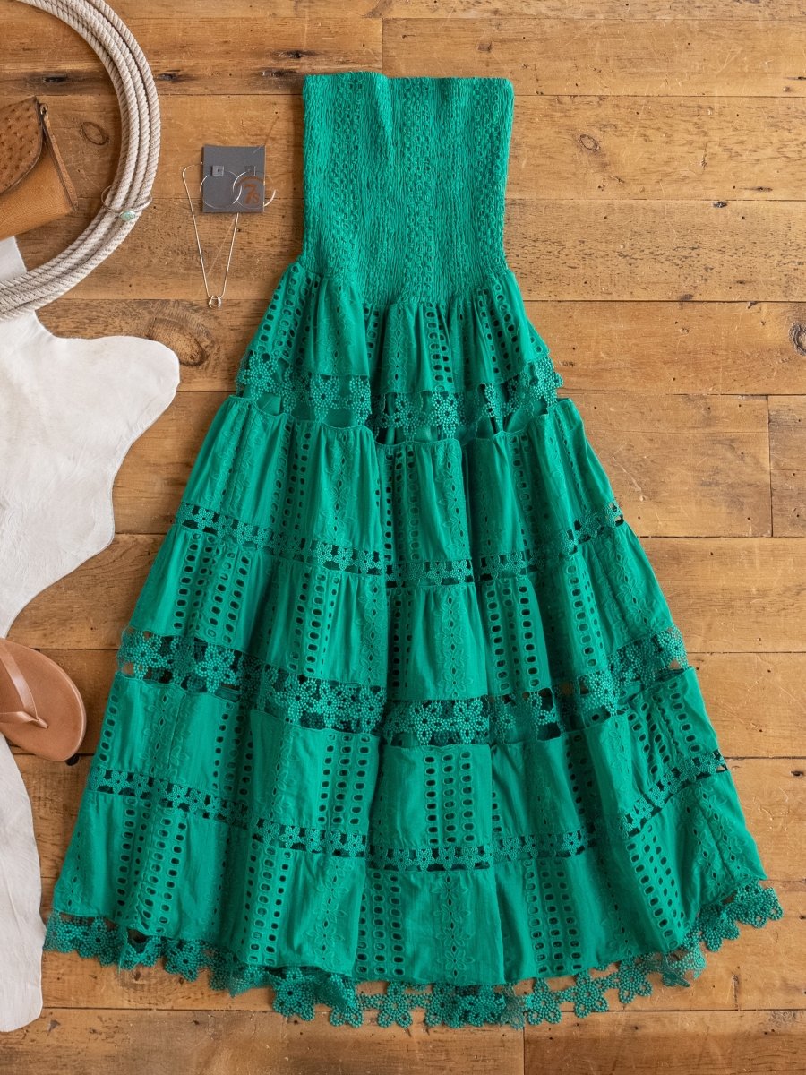 The Tekoa Jade Teal Crochet Strapless Dress by Savannah Sevens western life{&}style features a smocked bodice and tiered skirt, shown laid flat on wood with jewelry, sandals, a rope bag, and a white cowhide rug for effortless style.