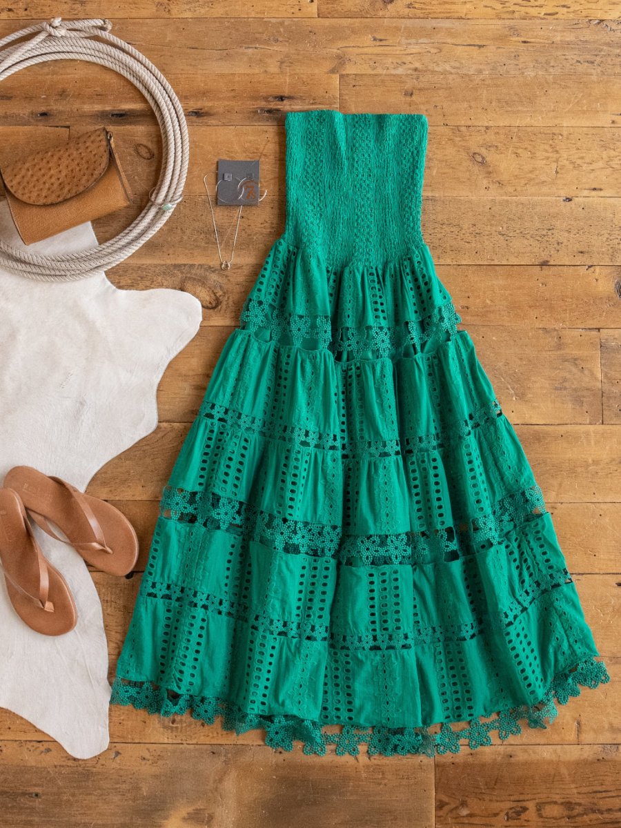 The Tekoa Jade Teal Crochet Strapless Dress by Savannah Sevens western life{&}style is displayed on a wooden floor with tan sandals, a tan handbag, and a silver necklace beside a white cowhide rug.