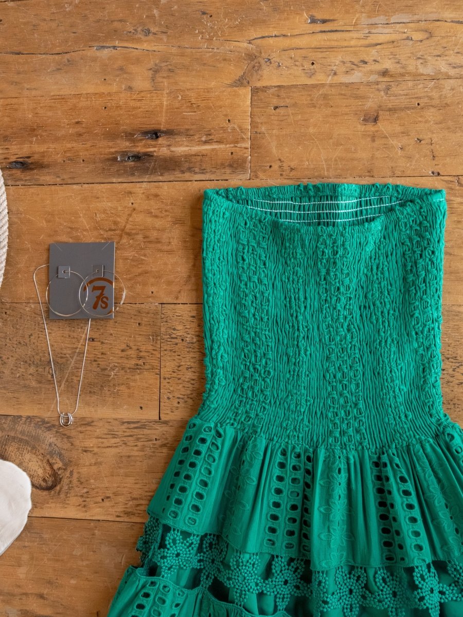 The Tekoa Jade Teal Crochet Strapless Dress by Savannah Sevens western life{&}style is displayed on a wooden floor beside a silver necklace and hoop earrings on a jewelry card.