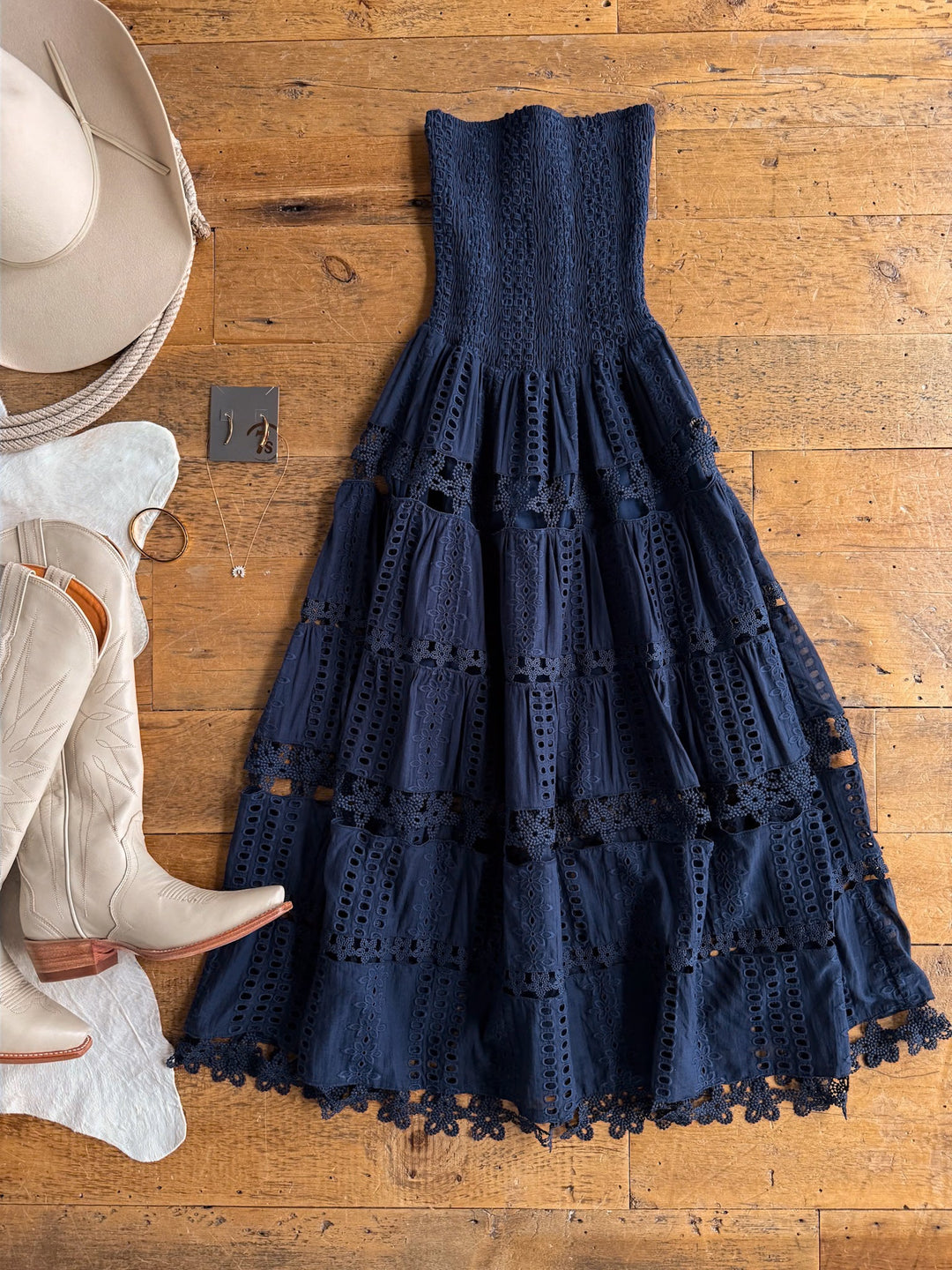 The Tekoa Navy Crochet Strapless Dress by Savannah Sevens western life{&}style is displayed on a wooden floor alongside a beige cowboy hat, matching cowboy boots, a necklace, and a pair of earrings.