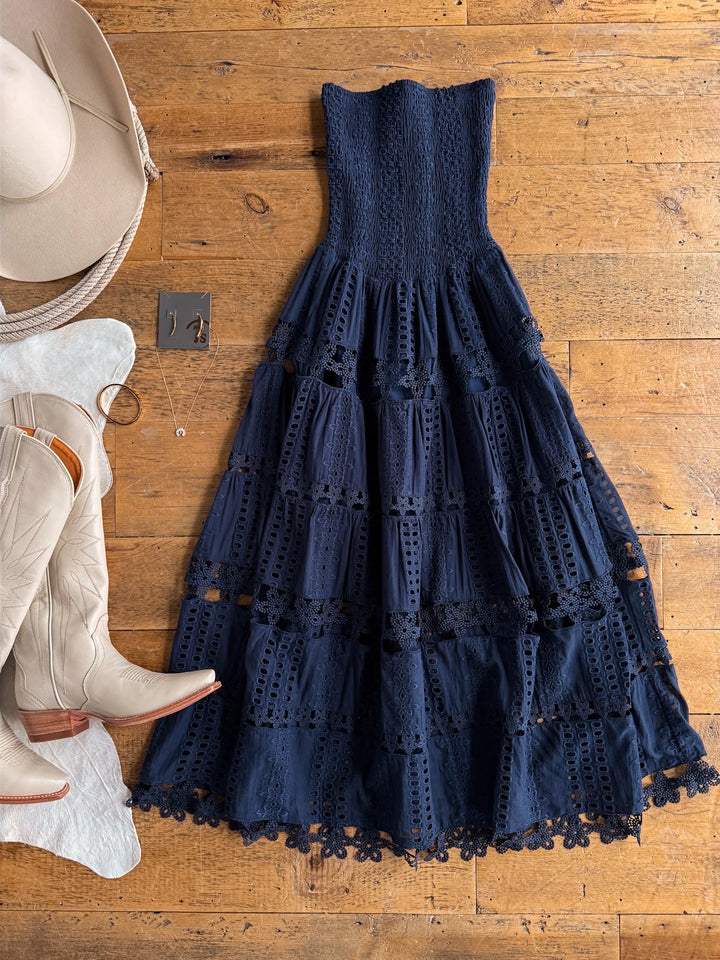 The Tekoa Navy Crochet Strapless Dress by Savannah Sevens western life{&}style is displayed on a wooden floor alongside a beige cowboy hat, matching cowboy boots, a necklace, and a pair of earrings.
